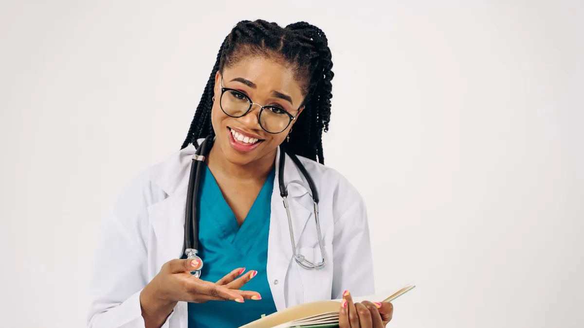 Smiling female doctor in a white coat, holding documents and wearing eyeglasses, seated and engaged.