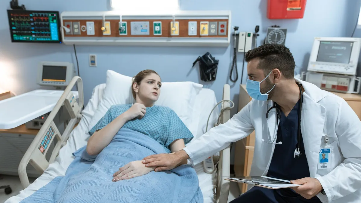 Male doctor with face mask comforts female patient in hospital bed with medical equipment.