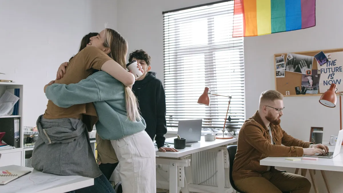 A group of diverse coworkers embrace and work on laptops under a rainbow flag.