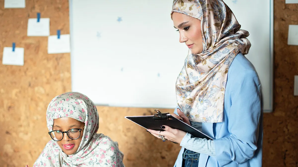 Two women wearing hijabs working together in an office with a laptop and clipboard.