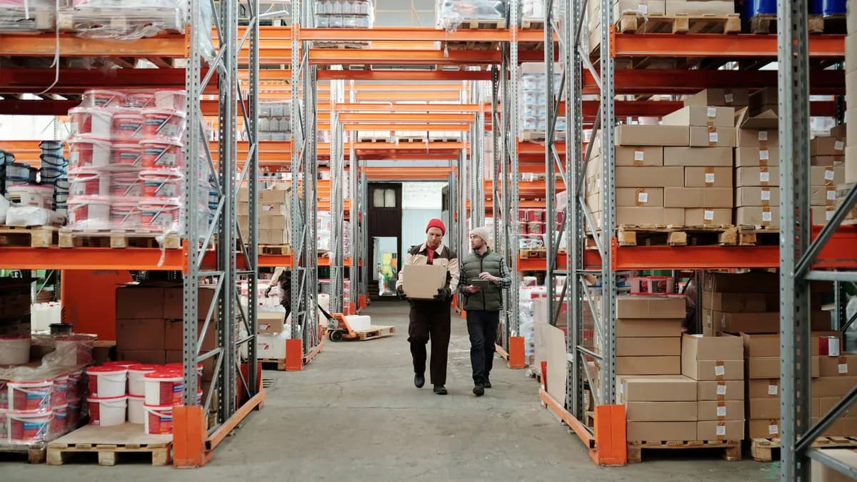 Two workers handle a package in a spacious warehouse surrounded by shelves stocked with boxes and products.