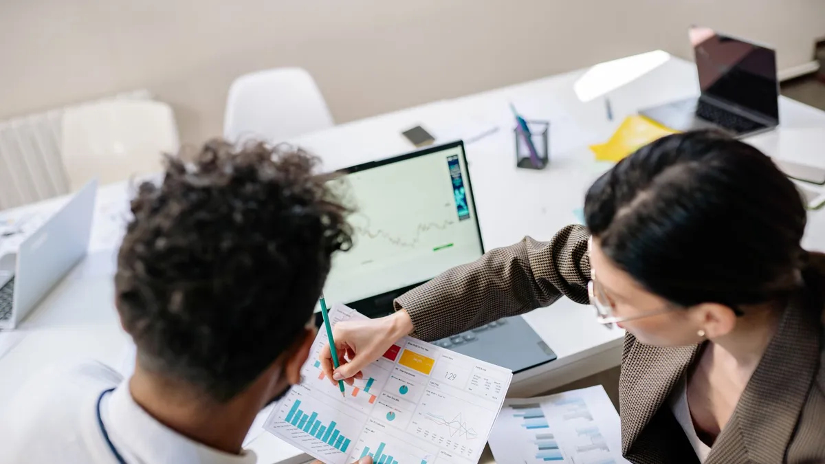 Two colleagues in a meeting room discussing financial charts and graphs on a laptop and paper.