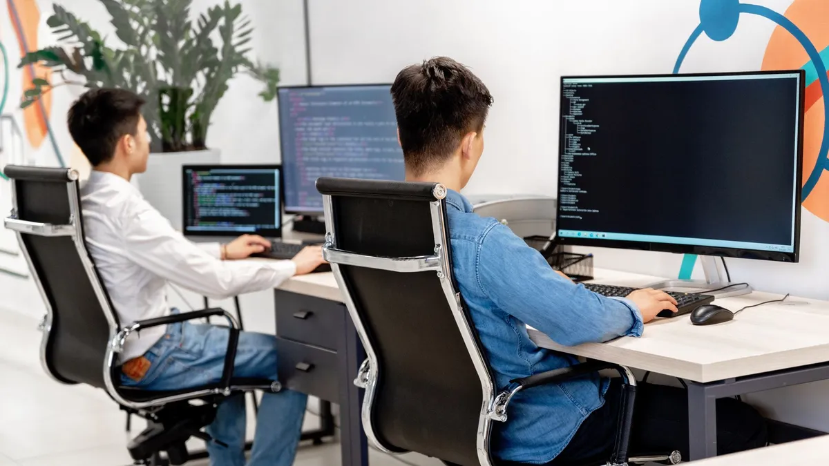 Two male developers at desks programming in a modern office workspace with large monitors.