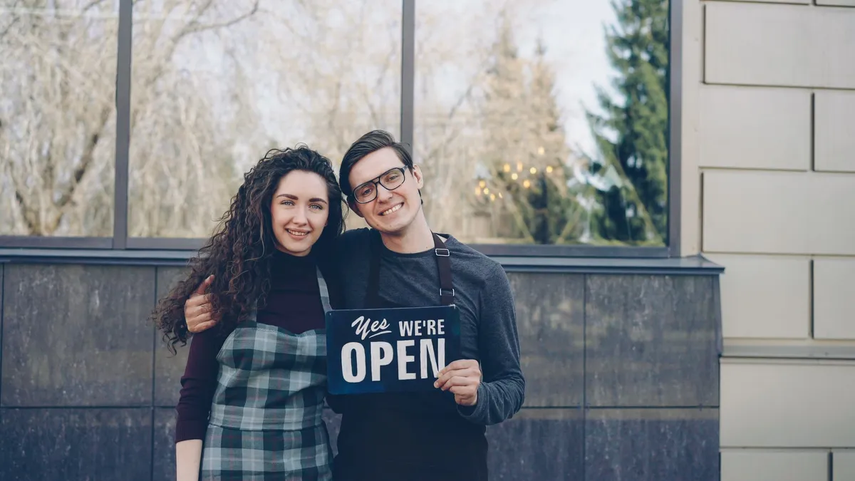 Happy small business owners embracing and displaying an open sign outdoors.